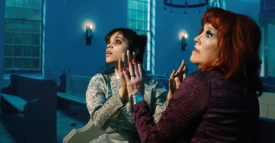 Jenna Ortega and Christina Ricci anxiously look upwards in a still from "Wednesday," in a scene set inside a dimly lit church