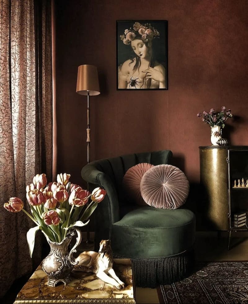 A green velvet chair in the corner of a room under a painting of a woman