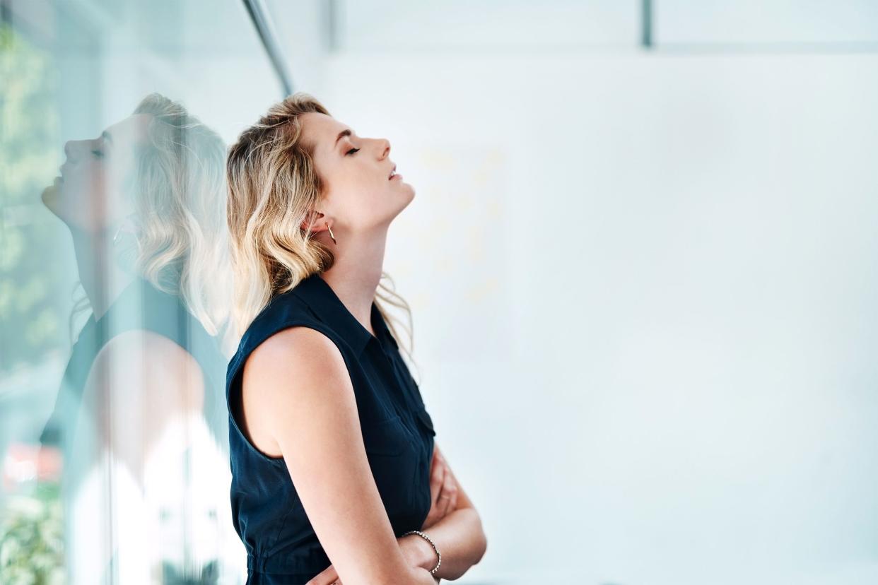 Side-view of stressed out woman leaning against a window with her head held up high, her arms are crossed, a white wall in the background and trees with leaves and bushes blurred behind the window