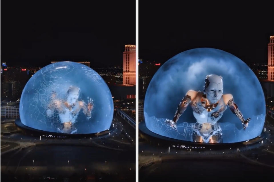 A large domed building displays a digital animation of a giant humanoid robot breaking glass, in a nighttime cityscape