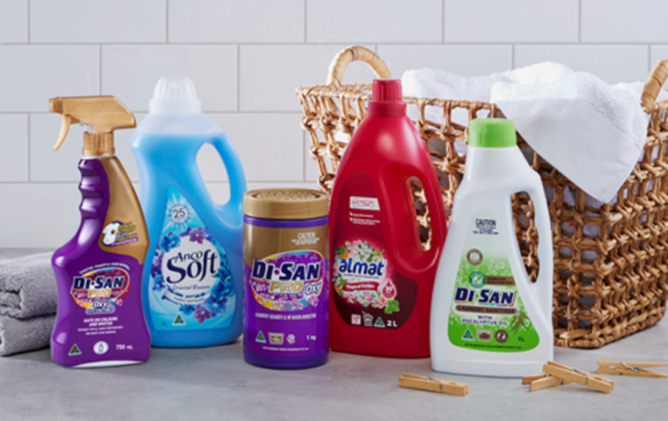 Five colourful bottles from the Aldi Di San range pictured with a wicker laundry basket, towel and pegs in a white tiled room.