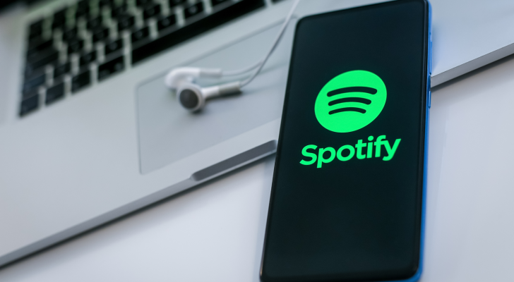 Close up view of a smartphone with Spotify (SPOT) logo on display. Laptop and headphone on background. New technology, social media, network, liquid music concept.