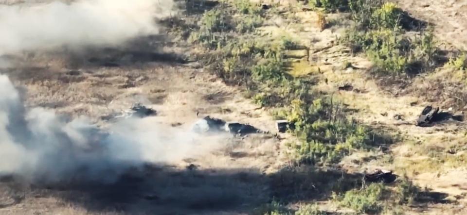 A still from drone video footage shows smoking tanks in a brown field.