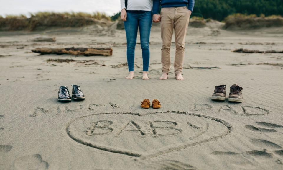 mum, dad and baby written in a heart on the sand of the beach next to their shoes with the legs of the parents behind