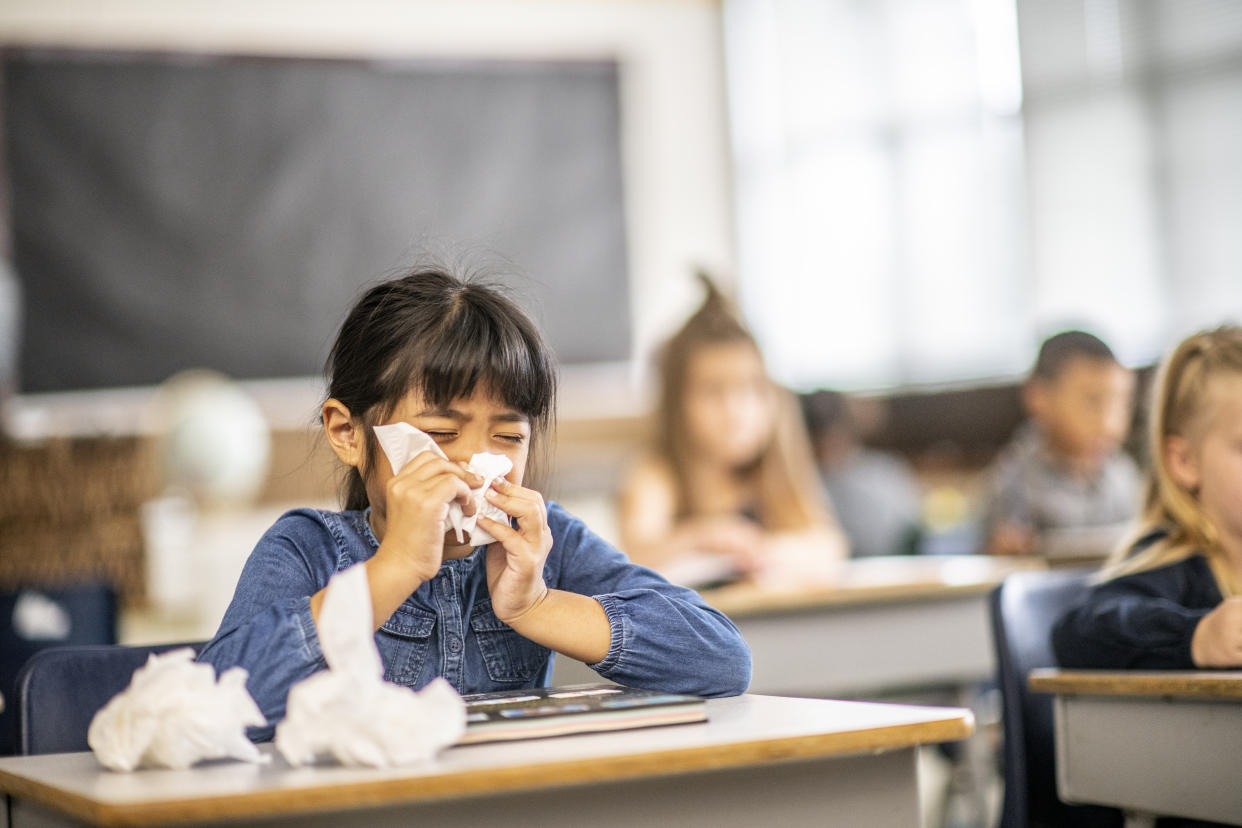 Young child blowing her nose with tissues at a desk in a classroom with other schoolchildren.