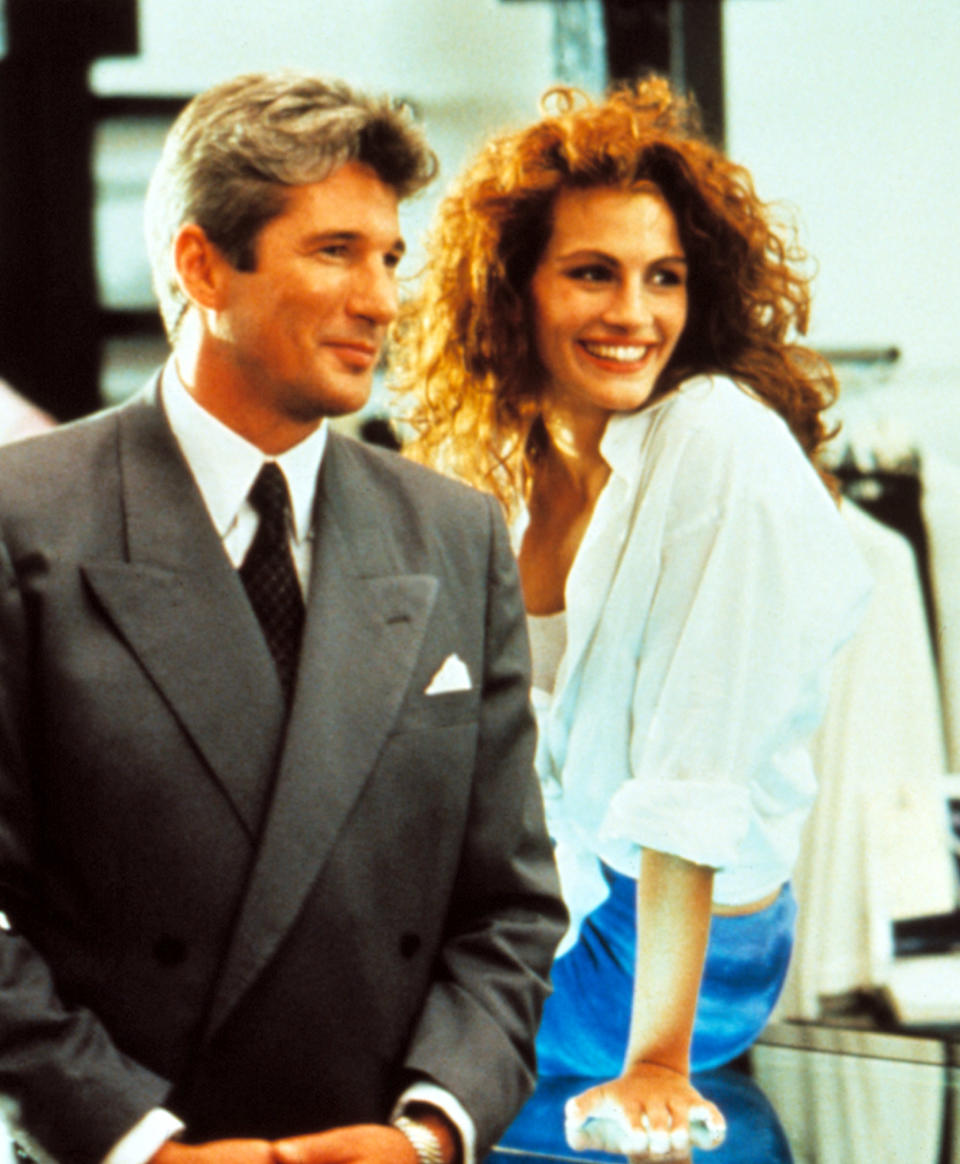 Richard Gere in a suit with his arms clasped in front of himself as Julia Roberts leans on a countertop next to him smiling widely in a scene from "Pretty Woman"