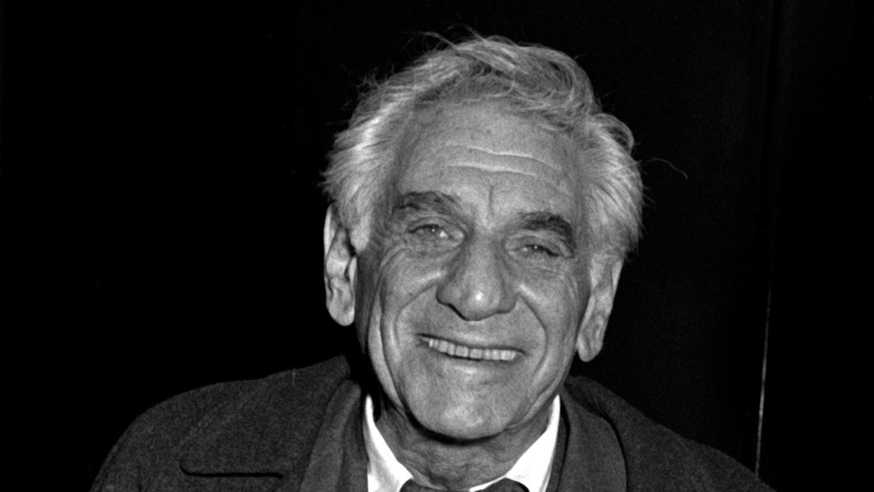 leonard bernstein smiles at the camera, he wears a wool coat and three piece suit with a white collared shirt and tie, he holds a pair of glasses in his right hand