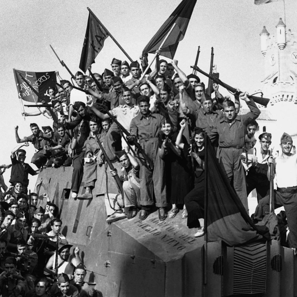 A large group of soldiers and civilians celebrate together on top of an armored vehicle, holding rifles and flags, during a historical military event