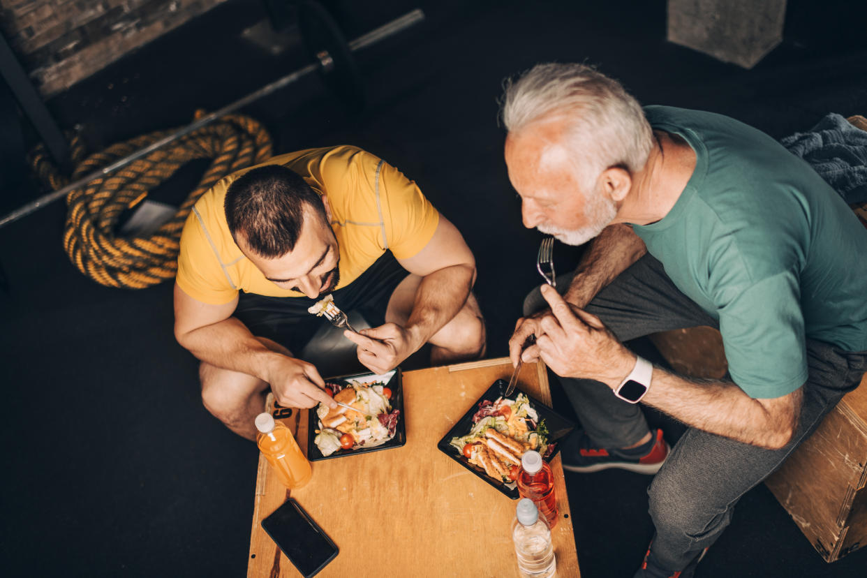 Active elderly sportsman eating a healthy protein chicken salad meal in a take-out box with his personal coach in a gym area of a gym.