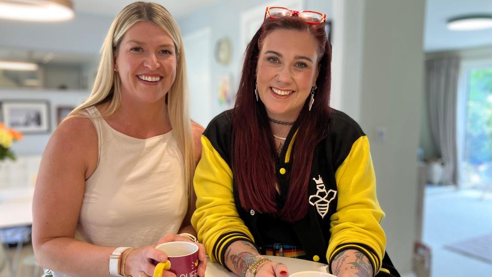 Emma Stirk and Dani Wallace smile for the camera and each hold mugs of tea in their hands 