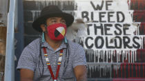 Native American advocate Carl Moore sits near the phrase "We Bleed These Colors" along a walkway which leads from the Bountiful High School parking lot up to the football field Tuesday, July 28, 2020, in Bountiful, Utah. While advocates have made strides in getting Native American symbols and names changed in sports, they say there's still work to do mainly at the high school level, where mascots like Braves, Indians, Warriors, Chiefs and Redskins persist. (AP Photo/Rick Bowmer)