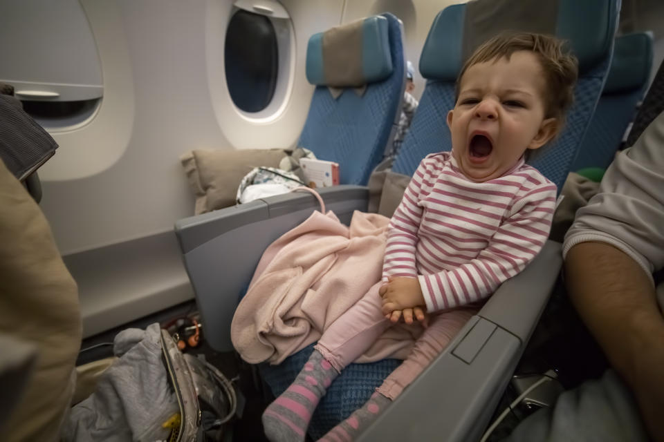 A toddler in a striped shirt yawns while sitting in an airplane seat. Blankets and a pillow are visible in the background