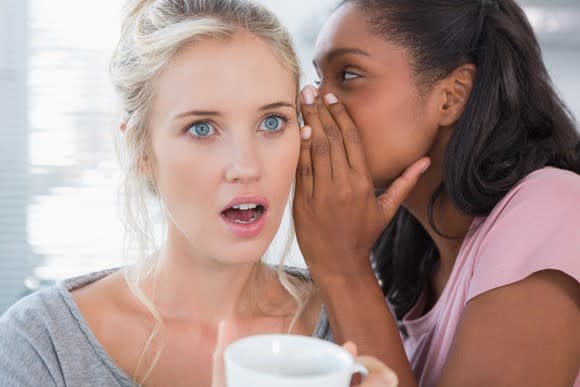 A young woman whispering to another young woman who is wide-eyed with her jaw dropped.