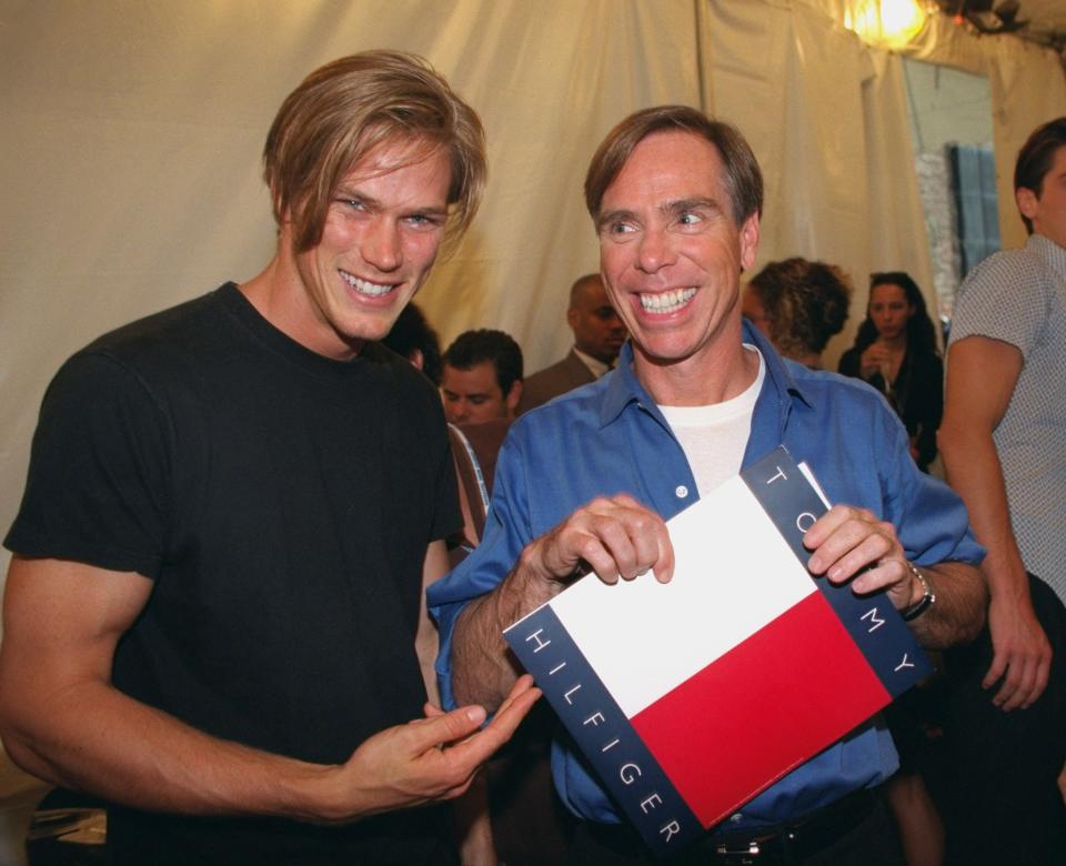 Sex and the City actor Jason Lewis stands in black t-shirt smiling and gesturing to designer Tommy Hilfiger with a big smile in blue shirt over white tee, fashion crowd in background