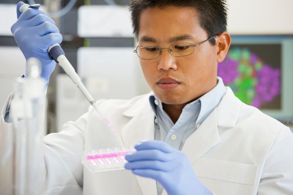 A lab technician wearing gloves using a pipette to place liquid samples into a row of test tubes.