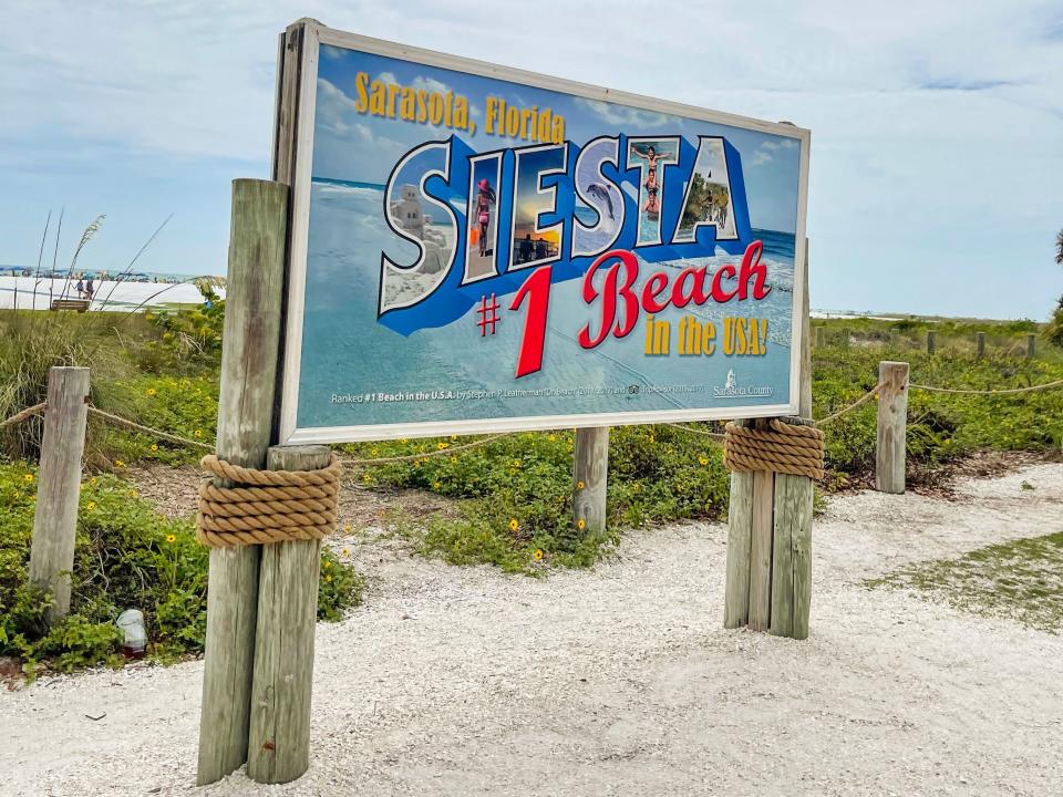 A blue sign with large block letters with images of people inside of them spelling out "Siesta" on a beach
