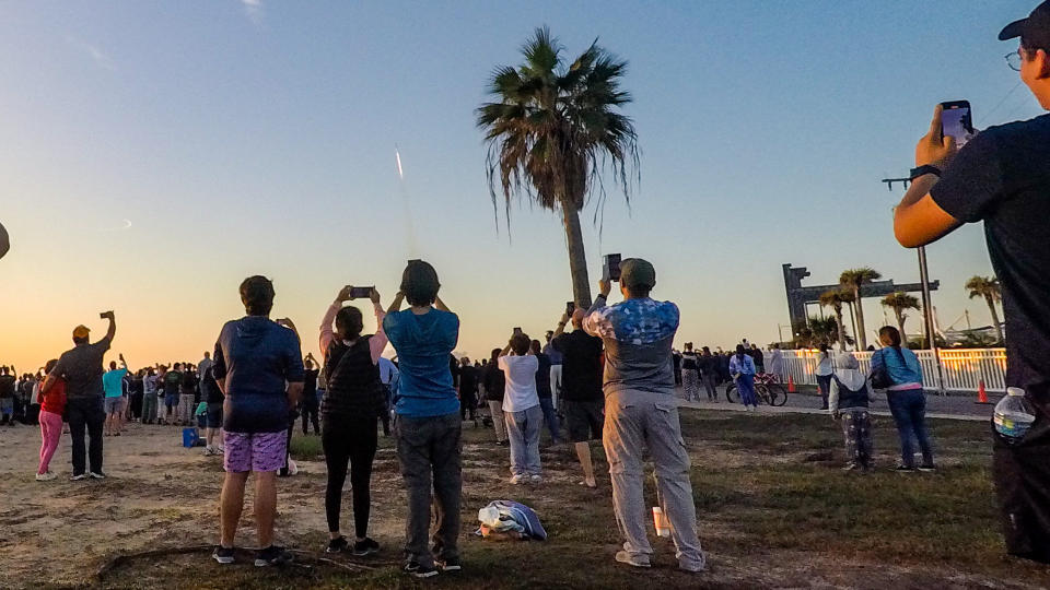 people look on as a roxcket launches, small in the distant sky.