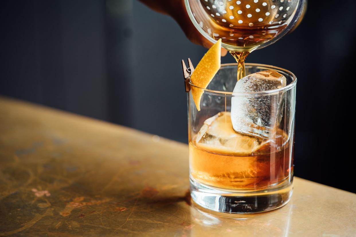 Focus on the tip of a cocktail shaker pouring negroni into a whiskey glass over ice with a slice of orange, on a copper table, with a blurred background of dark grey