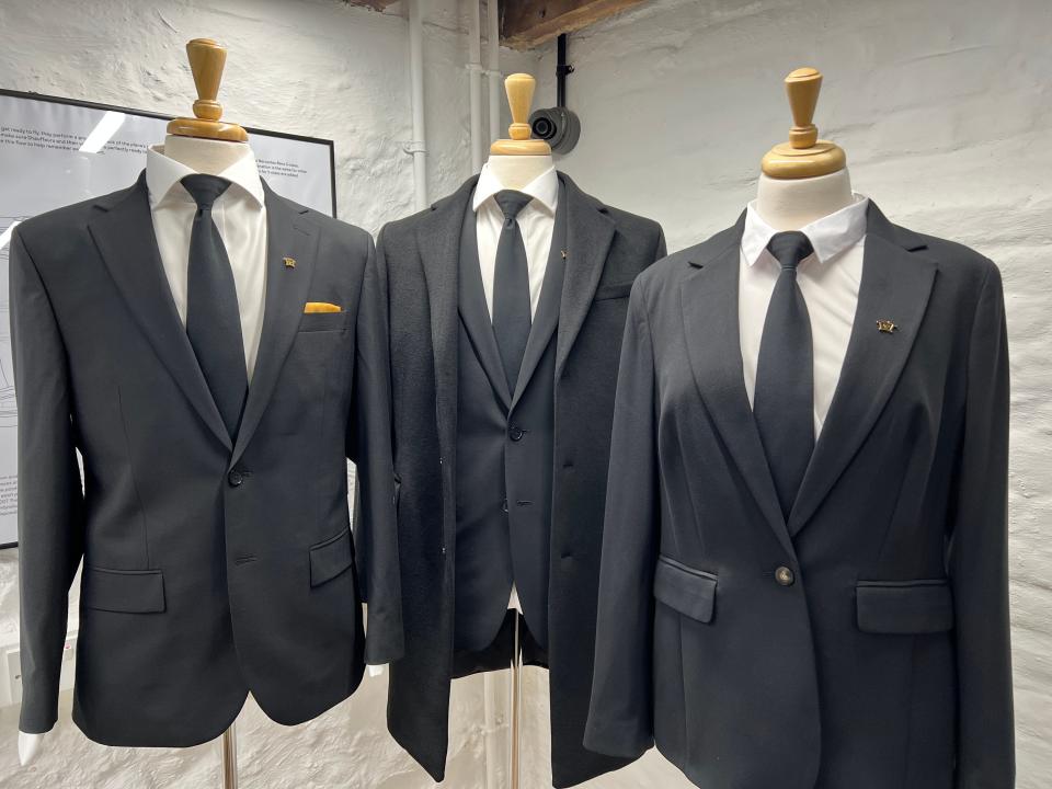 Three top-half mannequins dressed in black suit jackets and ties with gold pocket square, one has an overcoat, another is female