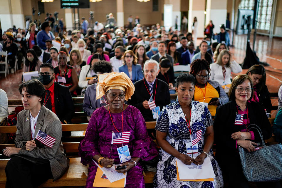 A woman wearing an African headdress and another in a batik outfit sit with other prospective citizens holding small American flags at a naturalization ceremony at Ellis Island.
