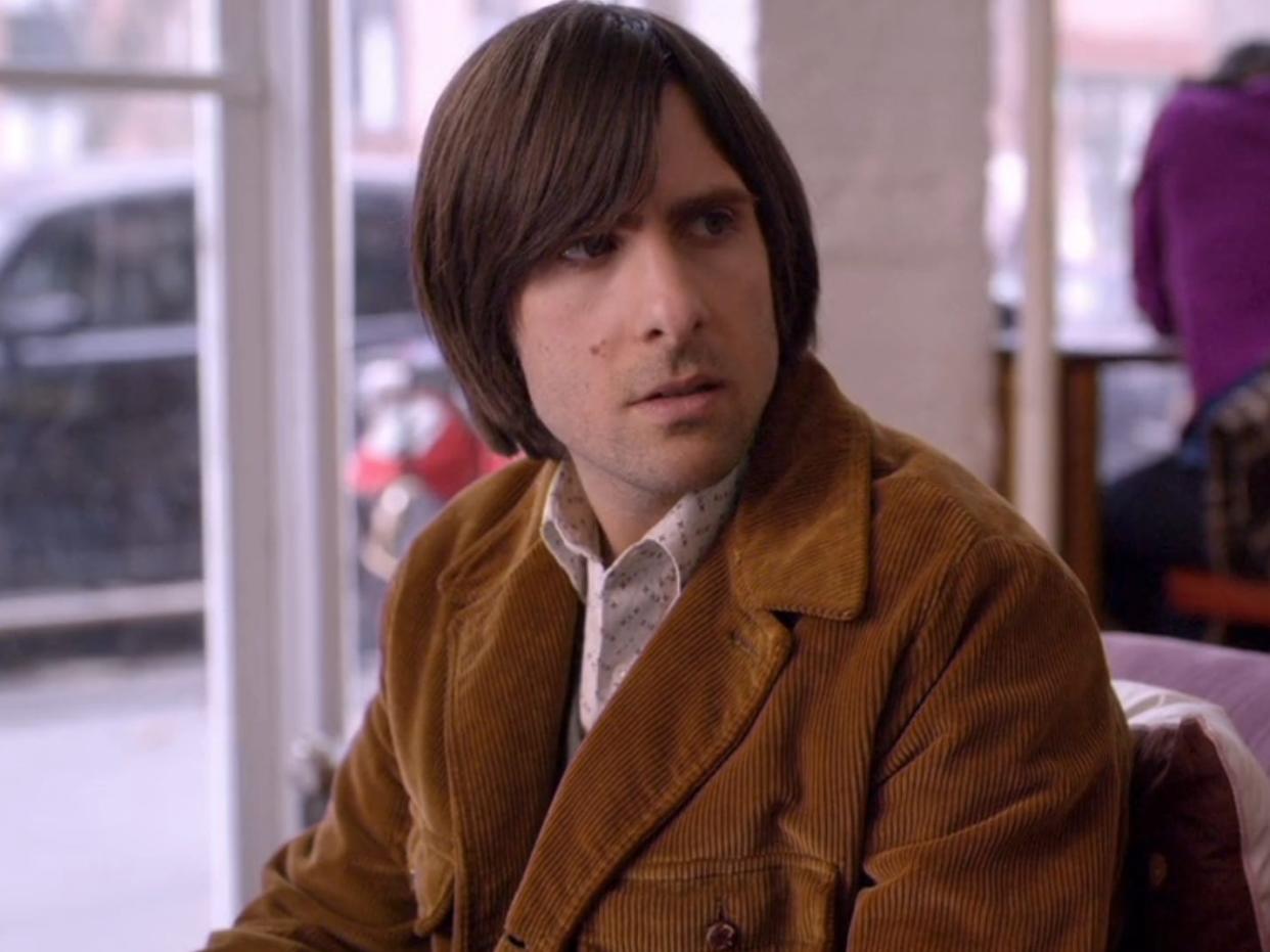 jason schwartzman as jonathan ames in bored to death. he's wearing a courduroy brown jacket, white buttom up, and his hair is worm jaw length and swooped to one side. he's sittingon a couch in a cafe