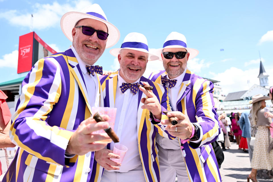 Three men smile for the camera and hold cigars while wear matching hats, bow ties and striped jackets.