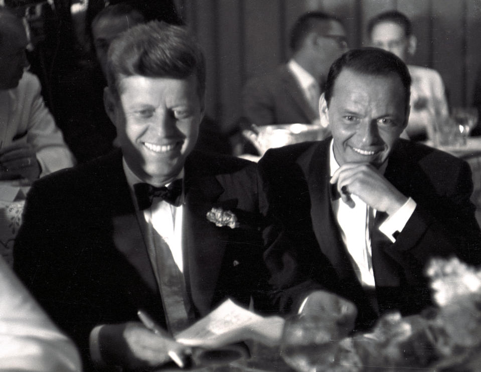 John F. Kennedy and Frank Sinatra sit together at a table, both smiling and dressed in formal suits. Other people in similar attire are in the background