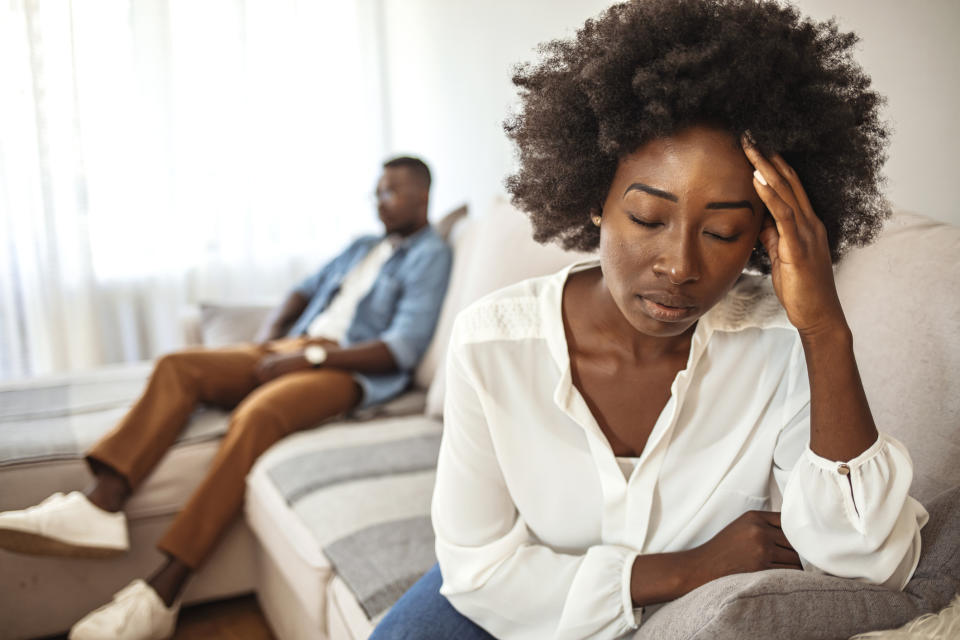 A woman in a white blouse sits on a couch looking distressed with her hand on her head. A man in a denim shirt and brown pants sits in the background