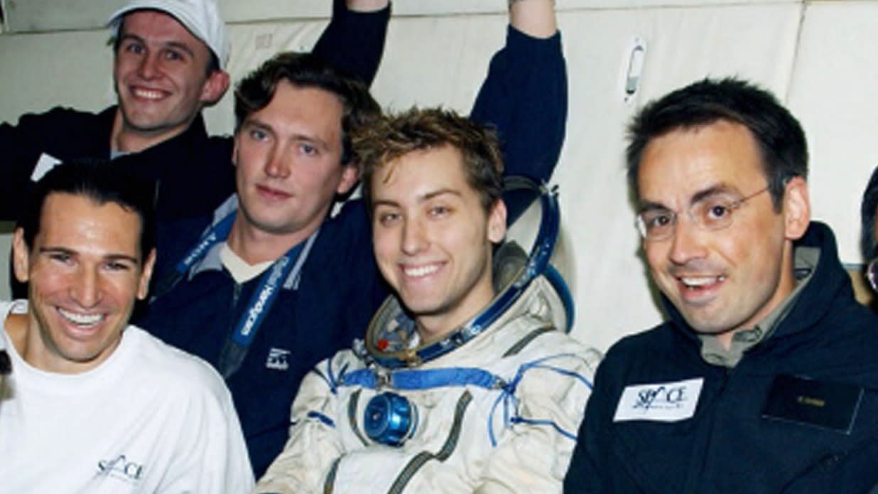  several people inside of a plane, sitting down. lance bass is at center, wearing a spacesuit and smiling 