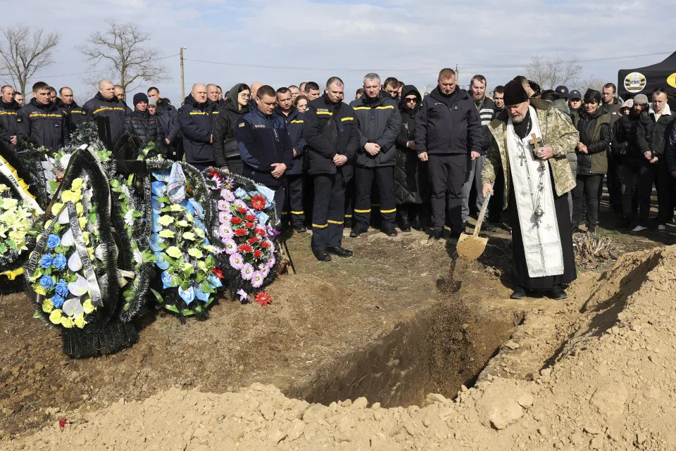 Emergency services workers look on as Military chaplain Archpriest Ioann shovels earth into the grave of Vitaliy Alimov during his funeral in Bilhorod-Dnistrovskyi, Ukraine, Monday March 18, 2024. Alimov, a firefighter, was killed in the Russian attack on Odesa on Friday March 15. (AP Photo/Victor Sajenko)