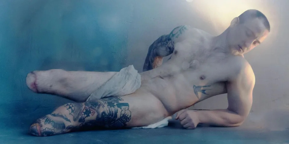 A photo by Marta Syrko of Ukrainian soldier Sasha, whose lower legs were amputated. Sasha, lying on his side and propped up by one elbow, is naked except for a strip of cream cloth over his loin, and looks down. He is muscular, has multiple tattoos and is bathed in a pearly, blue-and-cream light.