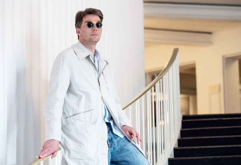 A man in a long jacket and small round sunglasses leans on the railing of a curving staircase in the foyer of an opera house