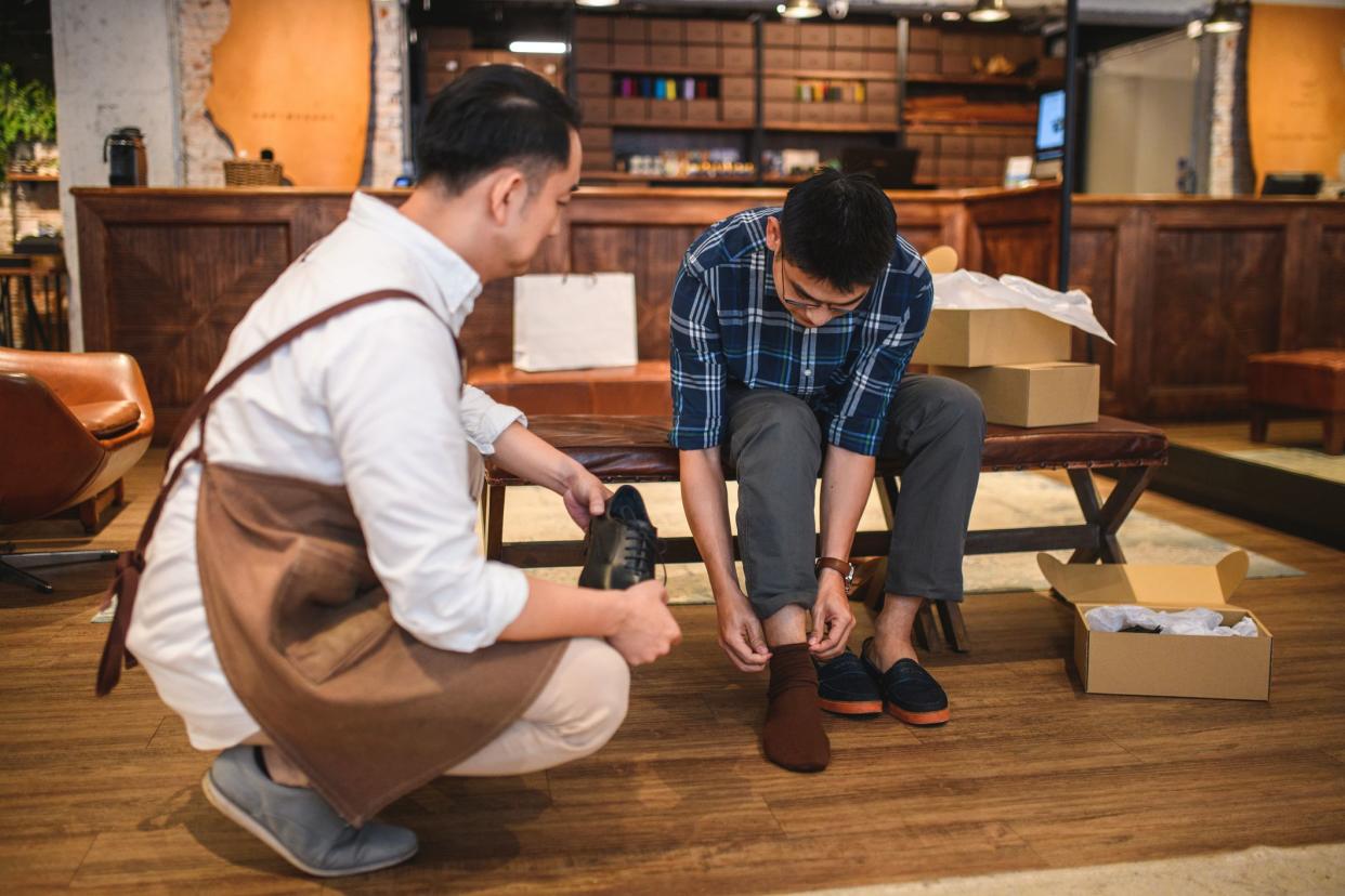 Young Korean male client sitting on a bench trying on formal leather shoes at a men's clothing store. A salesman is next to him holding a black leather shoe and helping him to try it on.