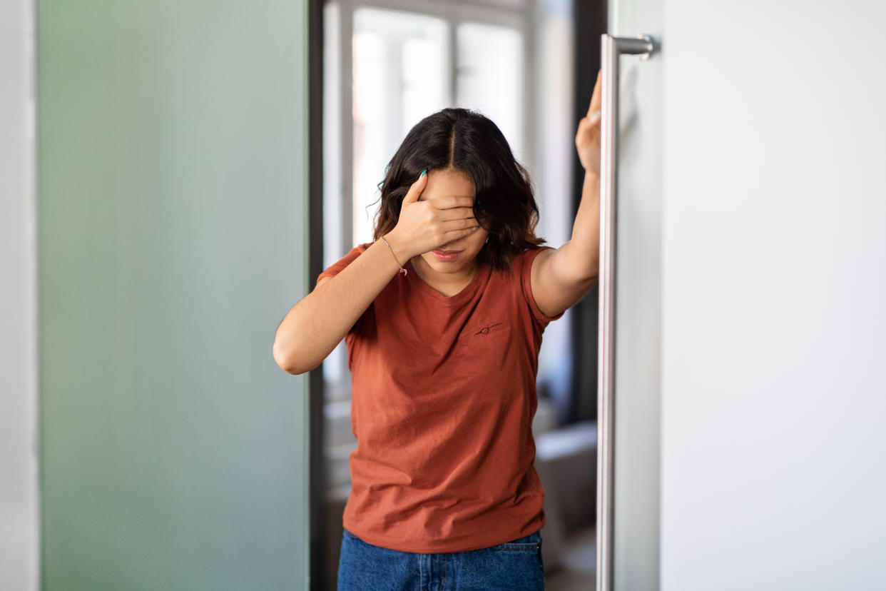 A woman stands in a doorway with one hand covering her eyes and the other pressed up against a wall.