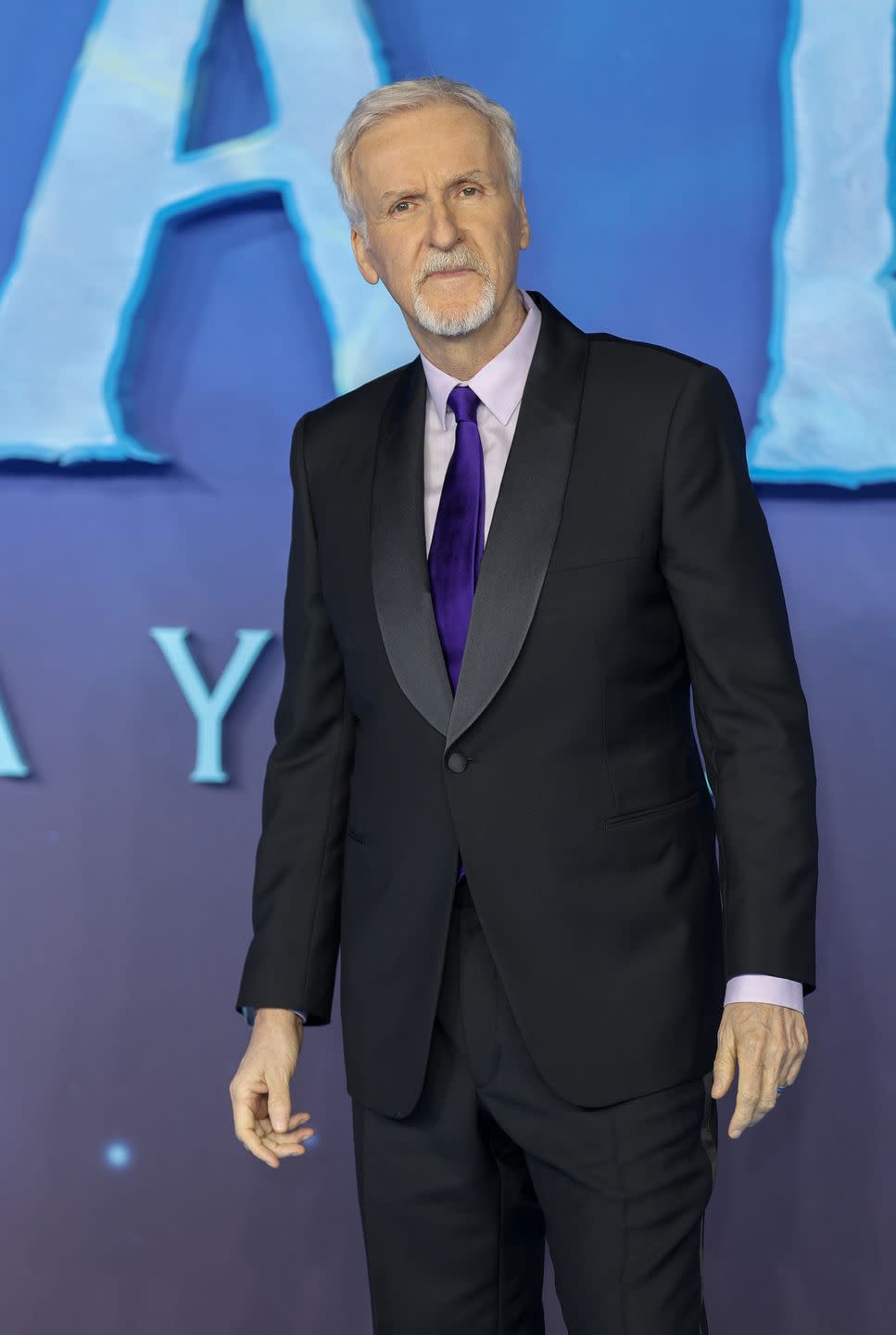 james cameron, an older man stands looking at the camera with a neutral facial expression, he wears a black suit with white shirt