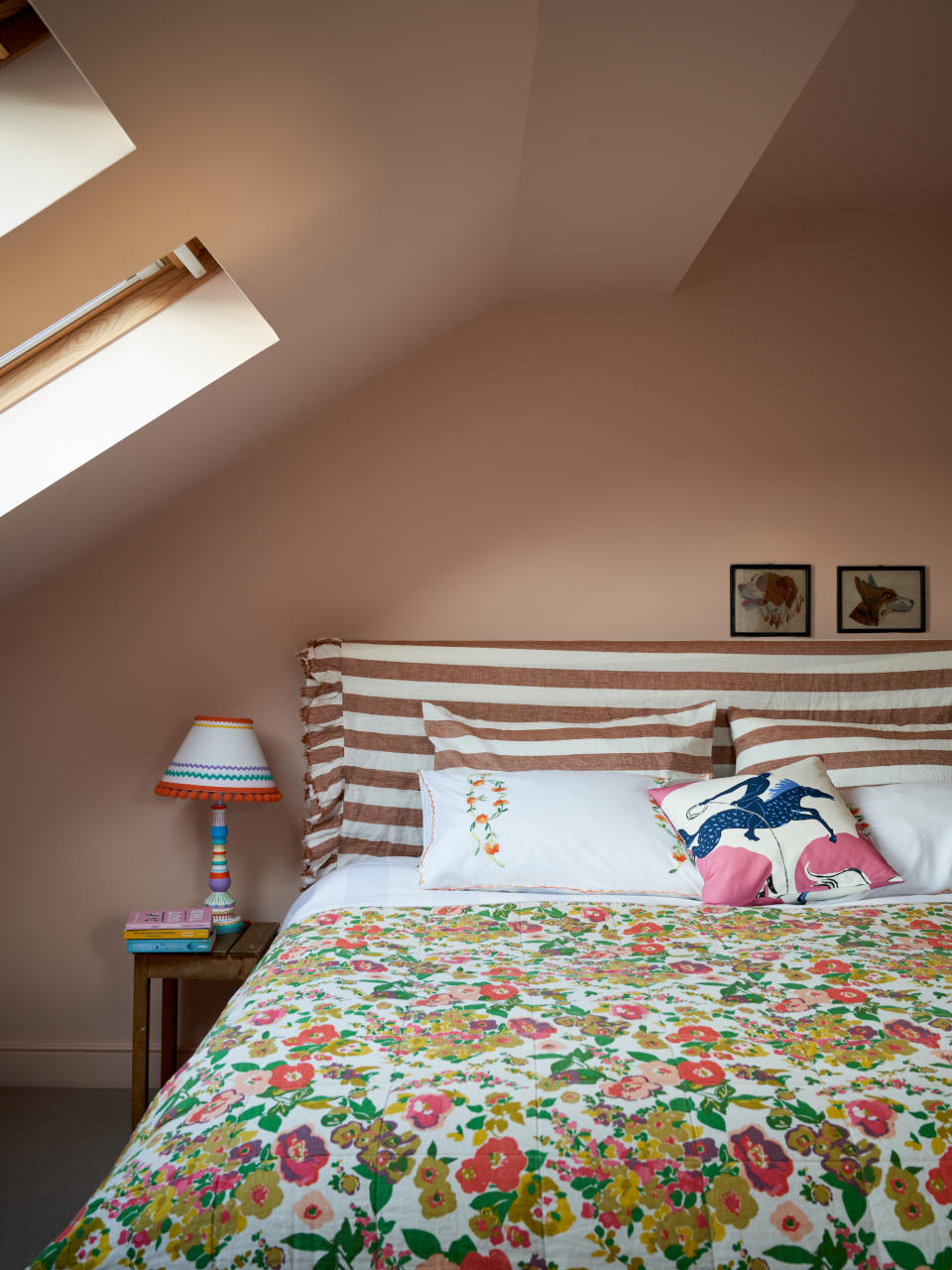A pink attic bedroom with a vaulted ceiling and two skylights housing a bed with striped headboard and floral bedding