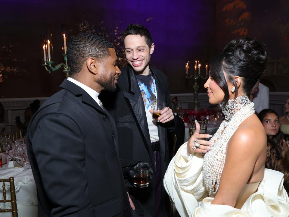 usher, pete davidson, and kim kardashian standing next to each other at the met gala. user is smiling, while davidson is grinning widely at him. across from usher, kardashian is smiling slightly with her mouth closed, her hand gesturing to her chest.