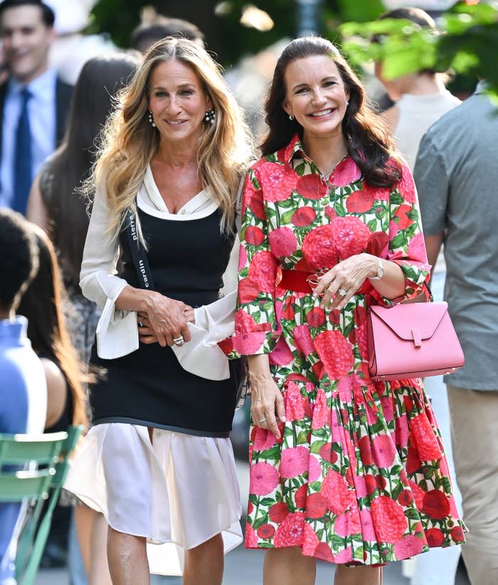 Sarah Jessica Parker and Kristin Davis walk outdoors. Parker wears a layered, monochrome outfit. Davis wears a floral dress with a pink belt and carries a matching bag