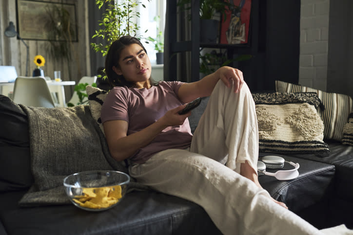 Person lounging on a sofa with a remote, bowl of chips nearby, in a cozy living room setting