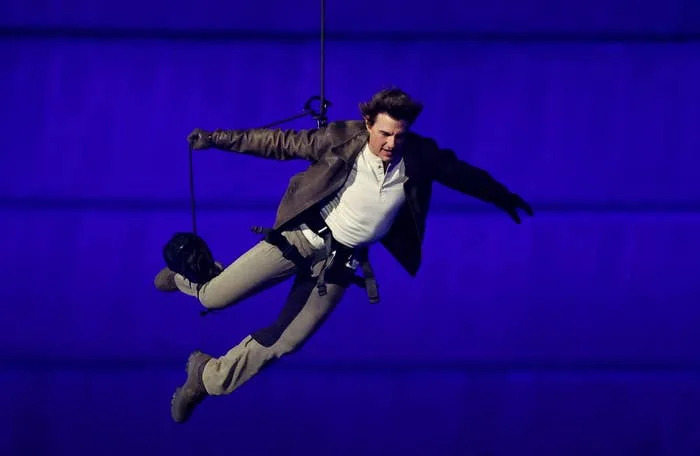 Tom Cruise performs a mid-air stunt dressed in a brown jacket and beige pants, suspended by cables against a blue backdrop