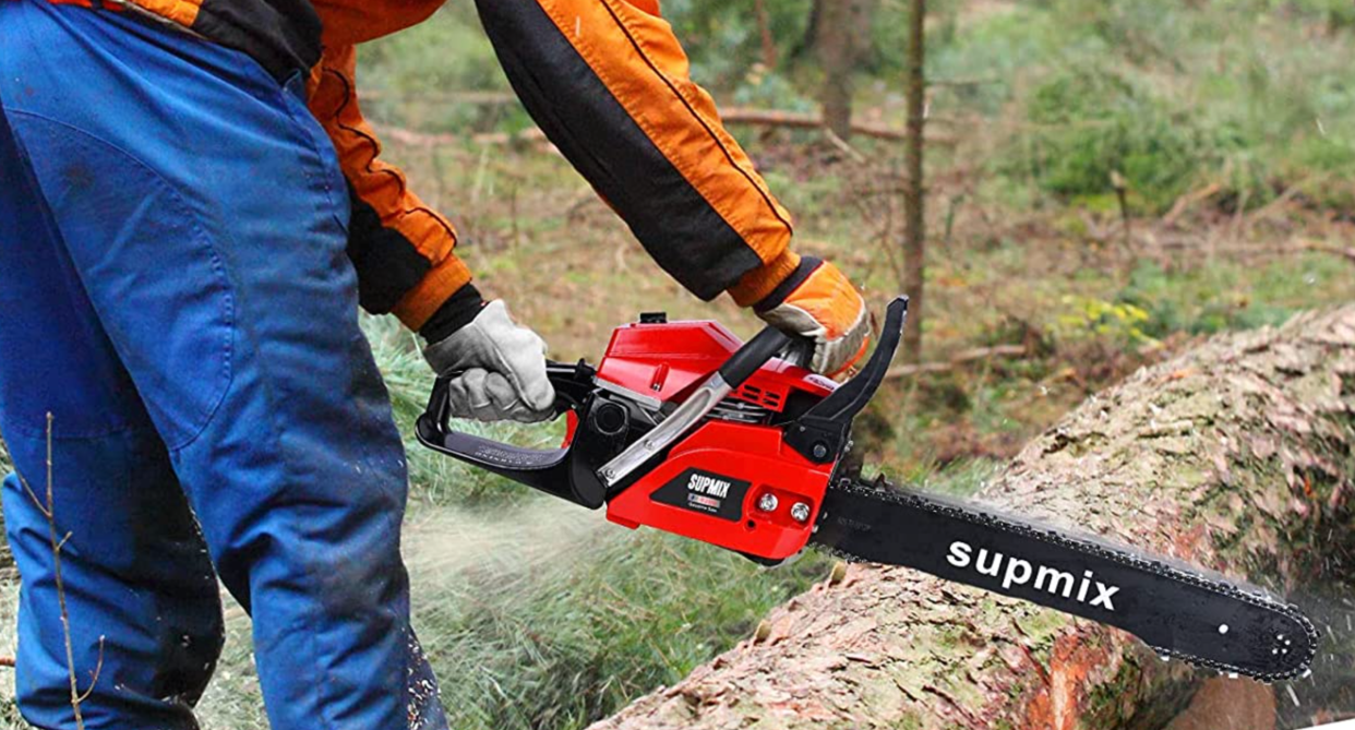 person wearing blue work pants, orange jacket holding red and black chainsaw cutting wooden log