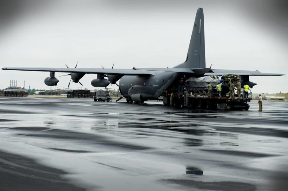 Workers in neon green safety vests and army fatigues stand on a platform while pushing cargo into the back of a large cargo plane.