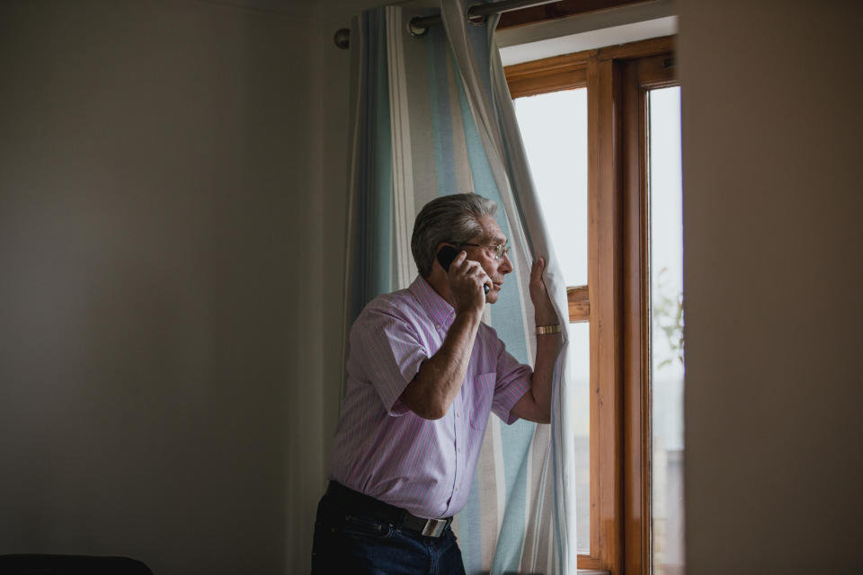 An elderly man in a striped shirt is talking on the phone while looking out of a window