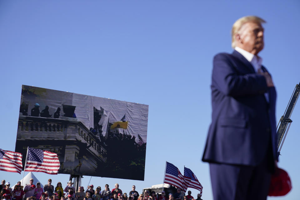 As footage from the Jan. 6, 2021, insurrection at the U.S. Capitol is displayed in the background, former President Donald Trump stands while a song, "Justice for All," is played during a campaign rally at Waco Regional Airport, Saturday, March 25, 2023, in Waco, Texas. The song features a choir of men imprisoned for their role in the Jan. 6, 2021, insurrection singing the national anthem and a recording of Trump reciting the Pledge of Allegiance. (AP Photo/Evan Vucci)