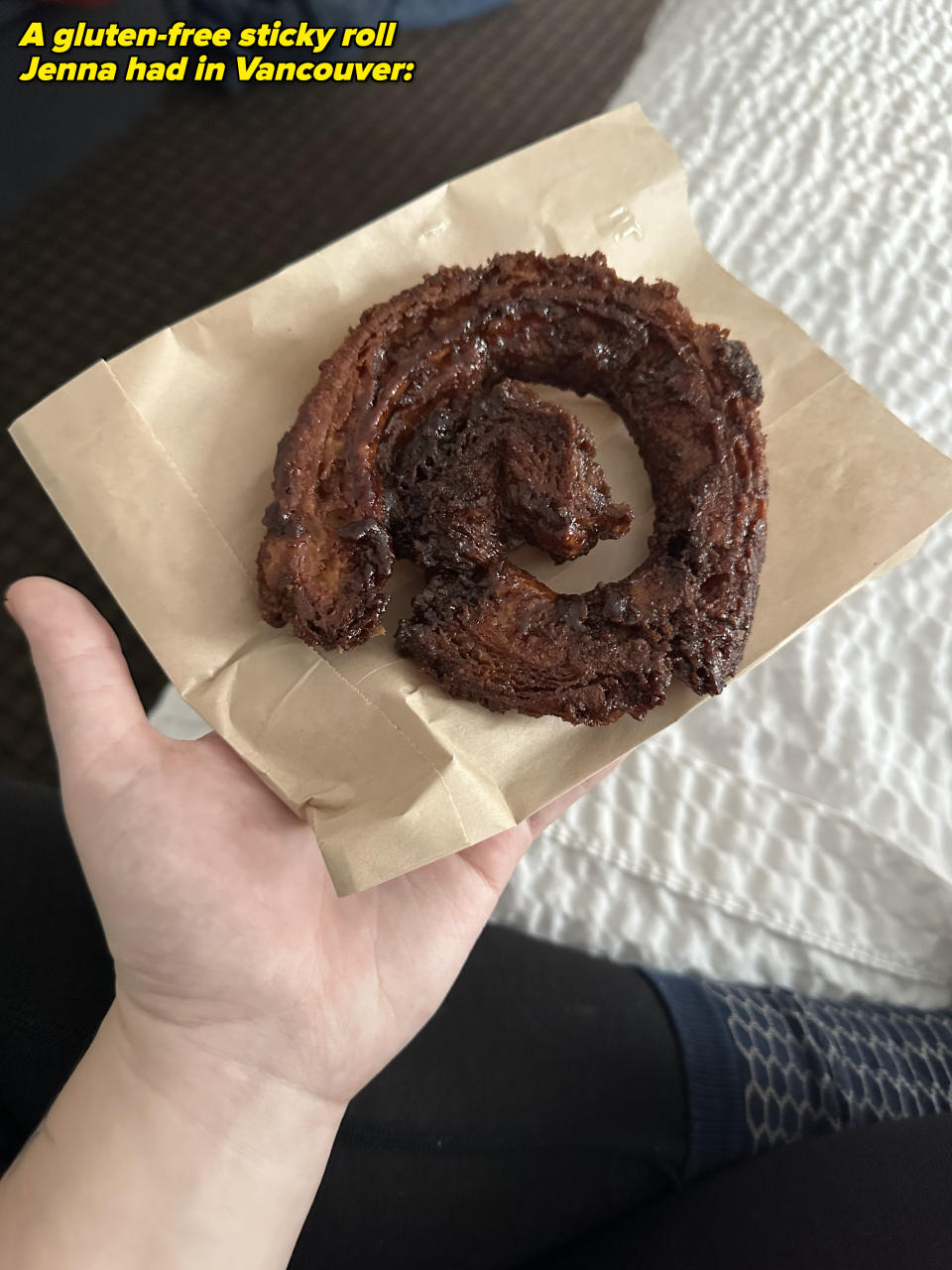 A hand holding a swirled, deep-fried pastry on a piece of brown paper, partially over a bedspread