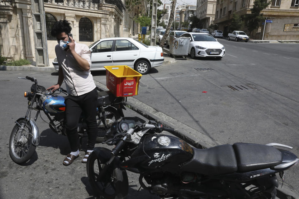 In this Tuesday, April 21, 2020 photo, grocery deliveryman Saeed Vatanparast, wearing a protective face mask to help prevent the spread of the coronavirus, speaks on his cellphone outside a store in Tehran, Iran. For some $15 a day, deliverymen don masks and gloves in Iran's capital to zip across its pandemic-subdued streets to drop off groceries and food for those sheltering at home from the virus. (AP Photo/Vahid Salemi)