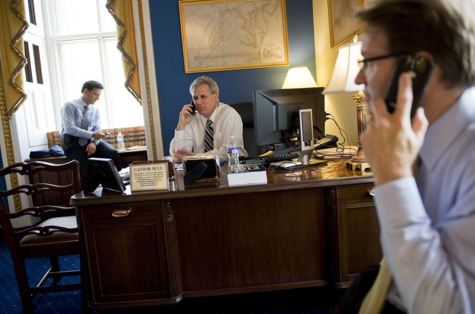 eric cantor, kevin mccarthy, and peter roskam makes call on their cell phones, they are sitting inside a congressional office at various seats, mccarthy sits in an office chair behind a large desk, he wears a white collared shirt and a black and yellow tie