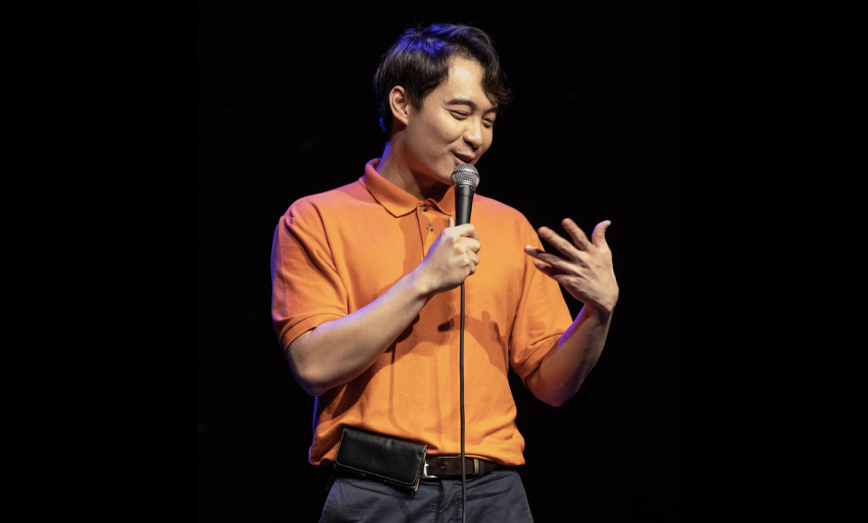 Nigel Ng performing as Uncle Roger in an orange polo tee in one of his stand up performances.