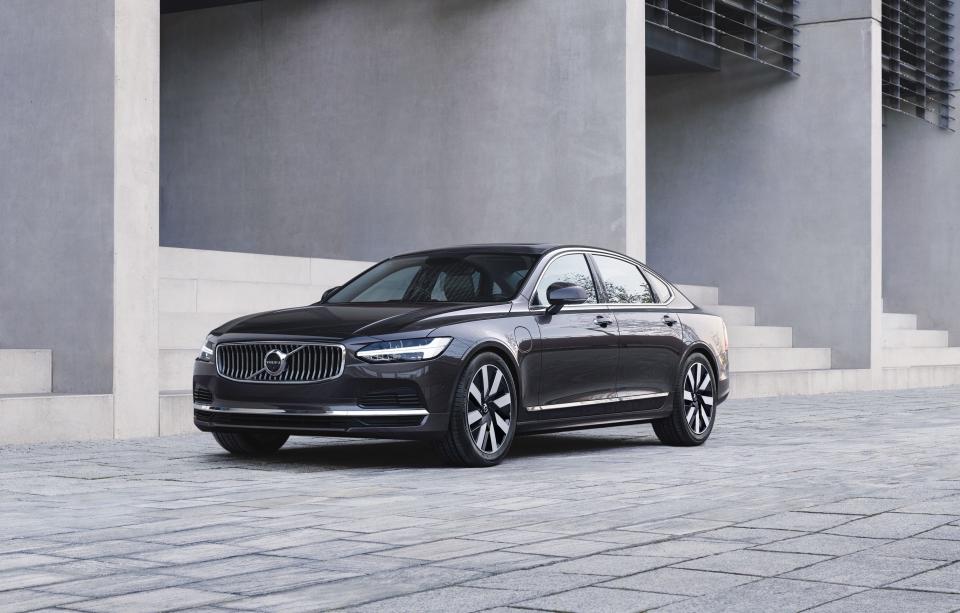 A gray Volvo S90 Recharge T8 AWD sedan is parked in front of a concrete building.