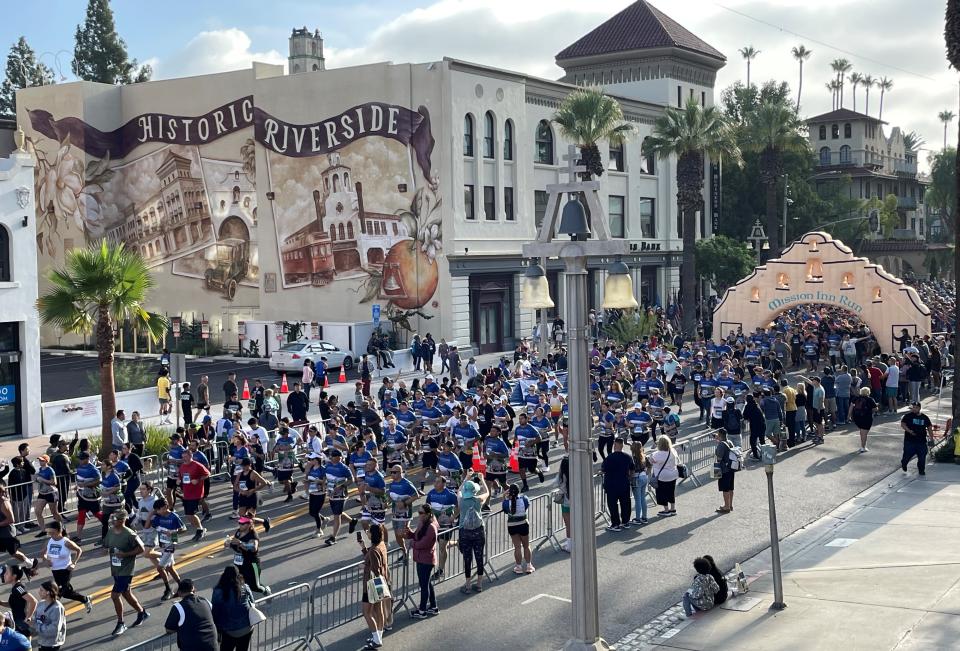 Marching band parades through a city street with onlookers, near buildings with 'Historic Riverside' mural